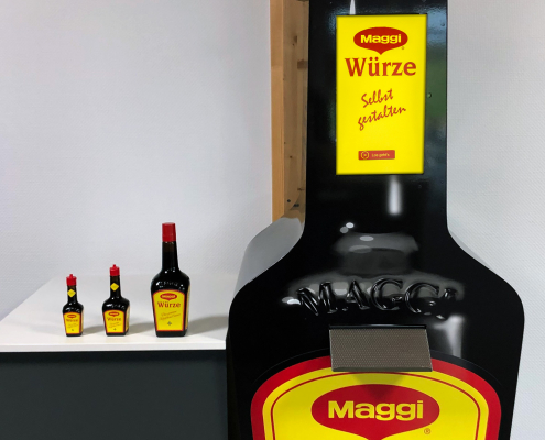 Lebensgroße Maggi-Würze Flasche mit drei normal großen Maggi Flaschen links daneben auf einem Tisch